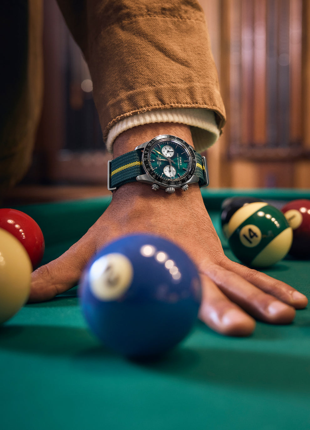 Lifestyle image of a model leaning over a billiard table while wearing the Timex Waterbury Heritage Chronograph, showcasing the green dial and strap in focus as colorful pool balls surround the wrist.