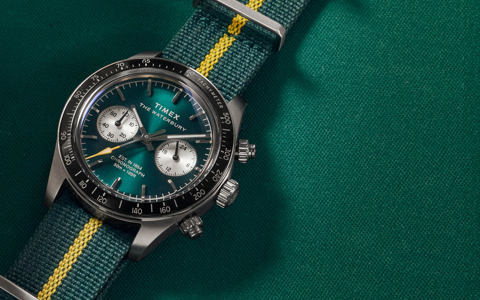 Close-up lifestyle image of the Timex Waterbury Heritage Chronograph featuring a deep emerald green dial, silver sub-dials, black tachymeter bezel, and a green fabric strap with a bright yellow center stripe, resting on a green billiard table surface.
