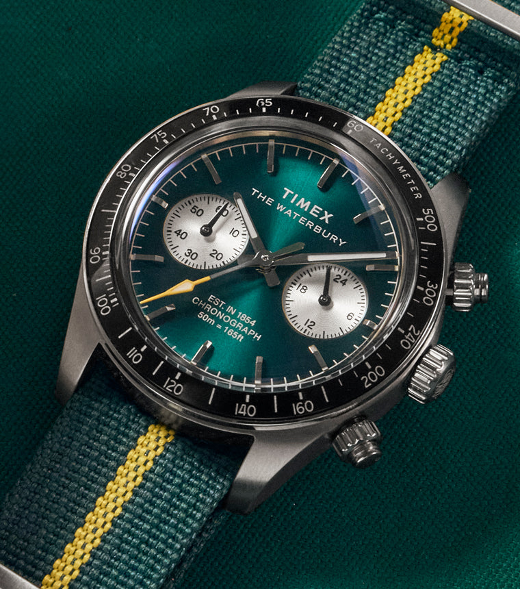 Close-up lifestyle image of the Timex Waterbury Heritage Chronograph featuring a deep emerald green dial, silver sub-dials, black tachymeter bezel, and a green fabric strap with a bright yellow center stripe, resting on a green billiard table surface.