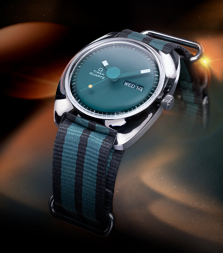 Stylized lifestyle image of the Q Timex® 1975 Enigma watch with a green dial and striped black-and-green fabric strap, illuminated by ambient orange and teal lighting against a cosmic-style background.