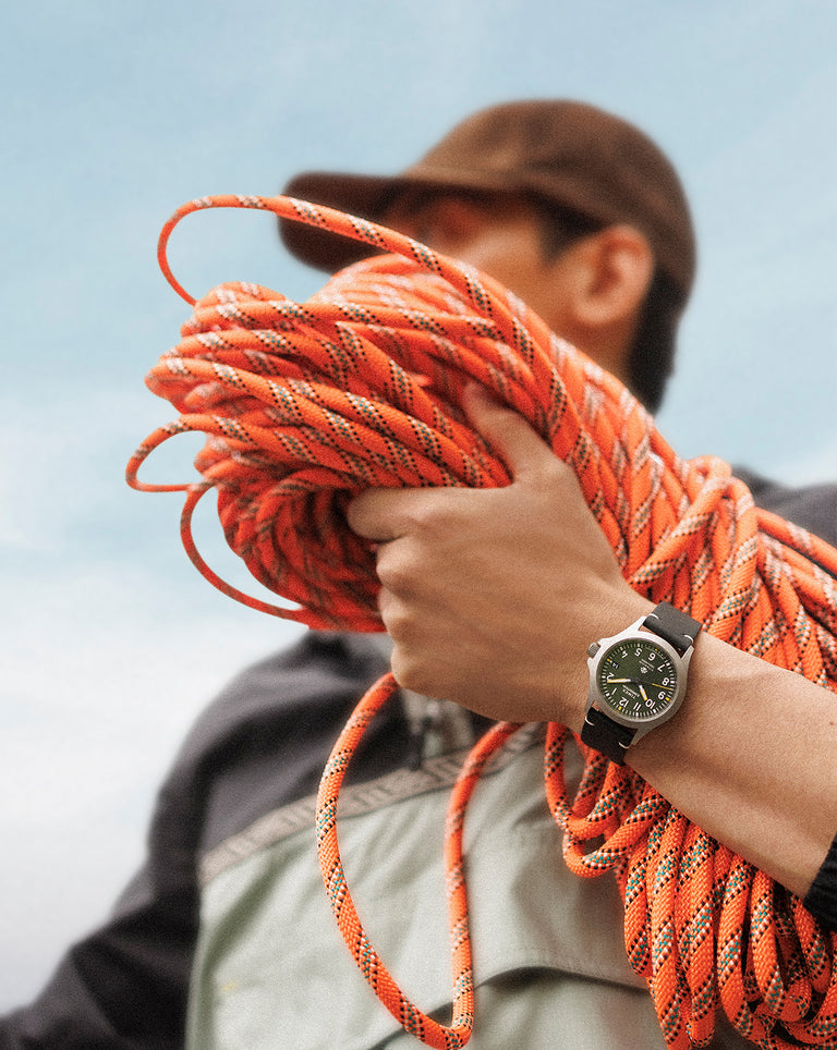 A man holds a bundle of bright orange climbing rope with one hand, wearing a rugged green dial field watch with a black strap. The scene conveys adventure and outdoor readiness, set against a clear sky.