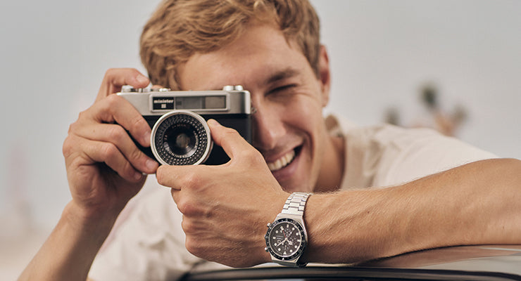 A cheerful man leans on a car while taking a photo with a vintage film camera. He wears a stainless steel chronograph watch and a white T-shirt, with a blurred beachside backdrop and palm trees in the distance.