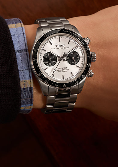 A Timex Waterbury Heritage Chronograph worn on a wrist, showcasing its stainless steel case and bracelet, black tachymeter bezel, and silver dial with black sub dials. The watch is styled with a dress shirt and jacket for a refined look.