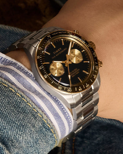 A Timex Waterbury Heritage Chronograph with a black dial and gold-tone subdials worn on the wrist, paired with a denim jacket and striped dress shirt, showcasing a bold yet classic casual look.