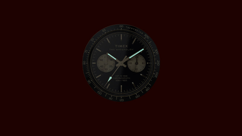 A view of the Timex Waterbury Chronograph dial in low light, showcasing its glowing luminescent hour and minute hands. The black tachymeter bezel and dual sub-dials remain subtly visible, emphasizing readability in the dark and the watch’s chronograph functionality.