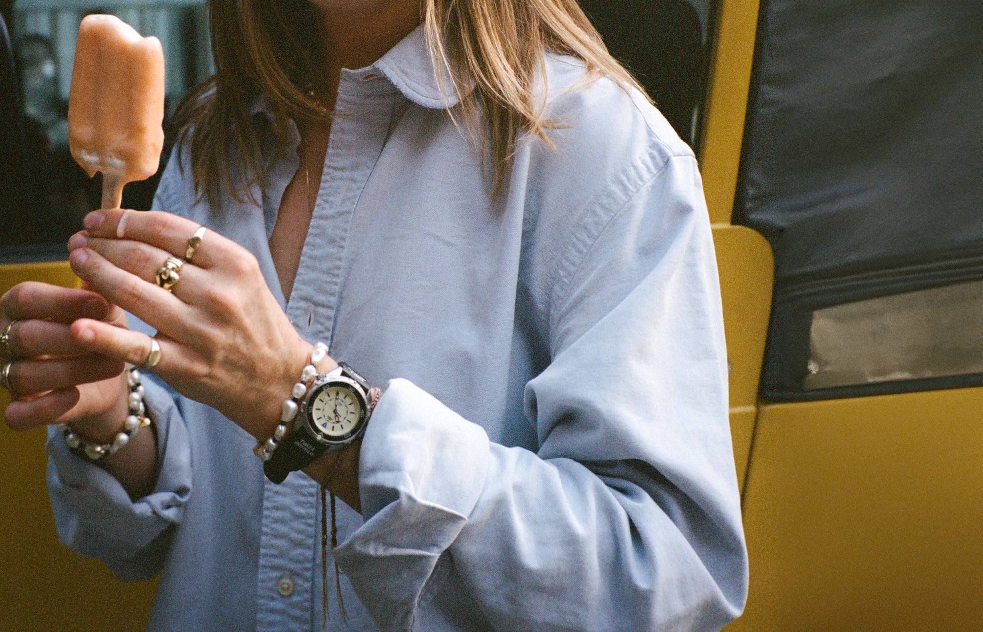 A close-up of a woman holding a partially eaten orange ice pop. She wears a light blue oversized shirt and multiple rings. Her wrist features the black Timex x Dimepiece watch with a pearlescent beaded bracelet beside it.