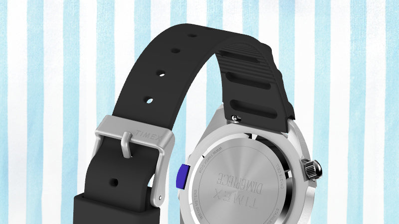 Digital render of the Timex x Dimepiece watch from a rear angle, showing the stainless steel case back engraved with the TIMEX and DIMEPIECE logos. The black rubber strap features a silver buckle engraved with “TIMEX” and a purple INDIGLO side button.
