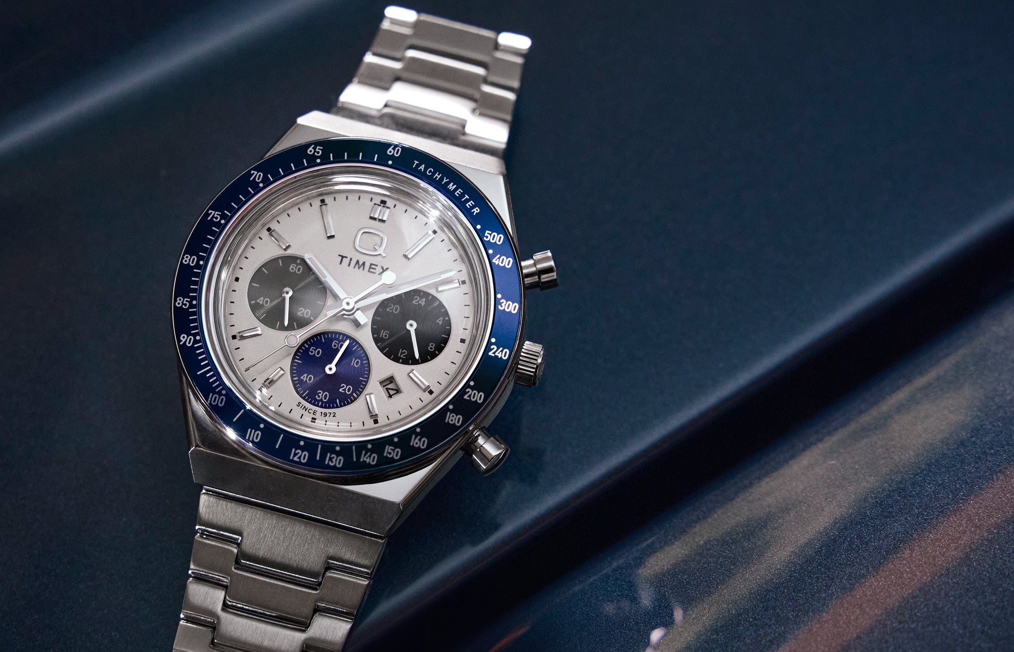 Q Timex® Chronograph on a metallic blue surface—white dial with three sub-dials, navy tachymeter bezel, date at 4 o’clock, stainless-steel bracelet.