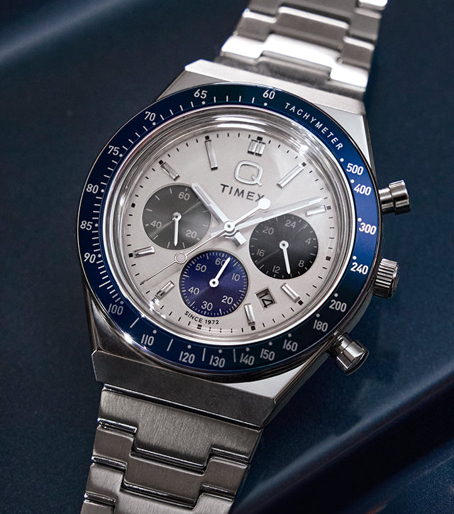 Q Timex® Chronograph on a metallic blue surface—white dial with three sub-dials, navy tachymeter bezel, date at 4 o’clock, stainless-steel bracelet.