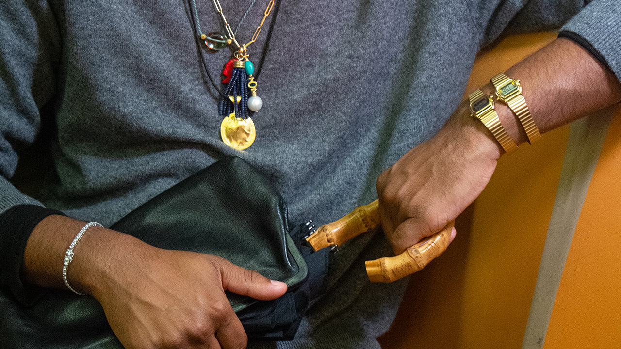 A person seated on a subway bench wearing two gold-tone Teeny Tiny Timex digital watches on one wrist. The watches complement layered gold necklaces and a bamboo-handled bag, reflecting Dimepiece’s nostalgic-meets-modern street style aesthetic.