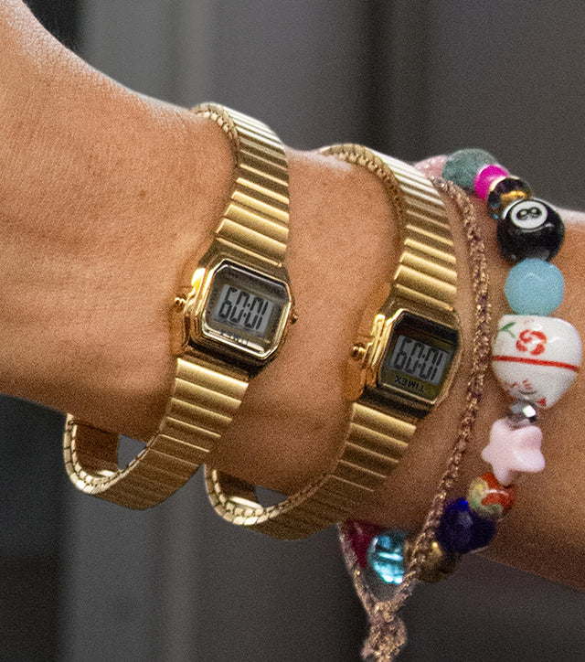 A person wearing two gold-tone Teeny Tiny Timex digital watches holds a yellow subway pole. The compact, retro-inspired watches are paired with colorful beaded bracelets and gold rings, capturing a playful and stylish city commute vibe.