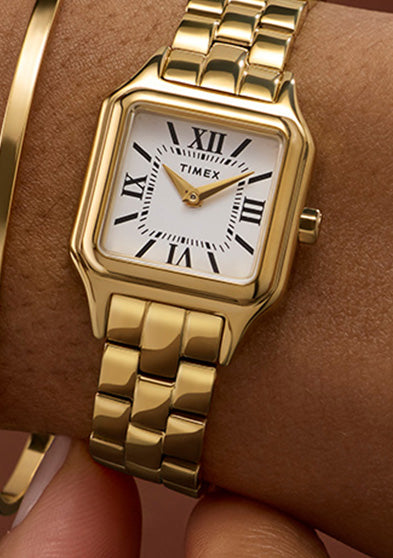 Timex Legacy Addison gold-tone rectangular watch with white Roman numeral dial, worn on the wrist beside a slim gold bangle.