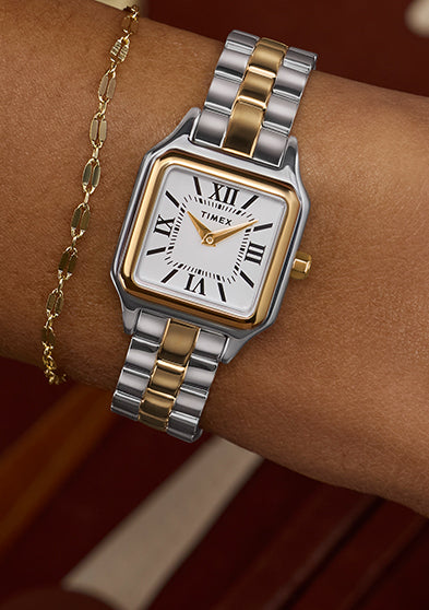 Timex Legacy Addison two-tone square watch with white Roman-numeral dial, worn on the wrist beside a delicate gold chain bracelet.