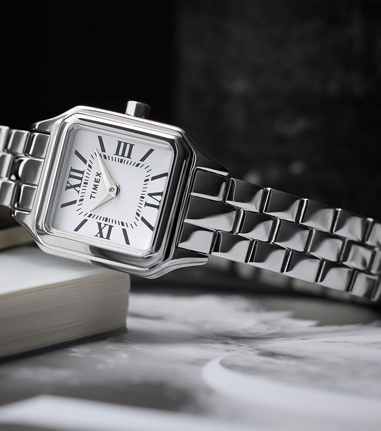 Timex Legacy Addison silver-tone women’s watch with square, rounded-corner case, white dial with black Roman numerals, and polished link bracelet, styled on books.