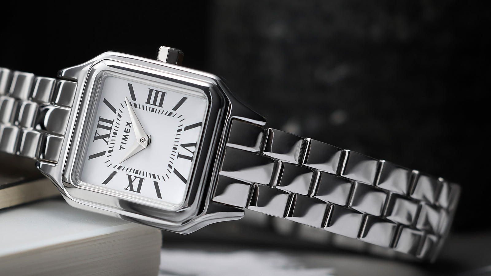 Timex Legacy Addison silver-tone women’s watch with square, rounded-corner case, white dial with black Roman numerals, and polished link bracelet, styled on books.