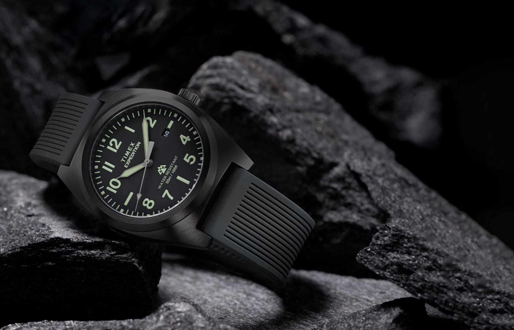 Timex Expedition® Capstone watch with black dial and black silicone strap resting among rugged dark rocks. The watch face glows softly with luminous markers, highlighting its durability, 50-meter water resistance, and adventure-ready design.