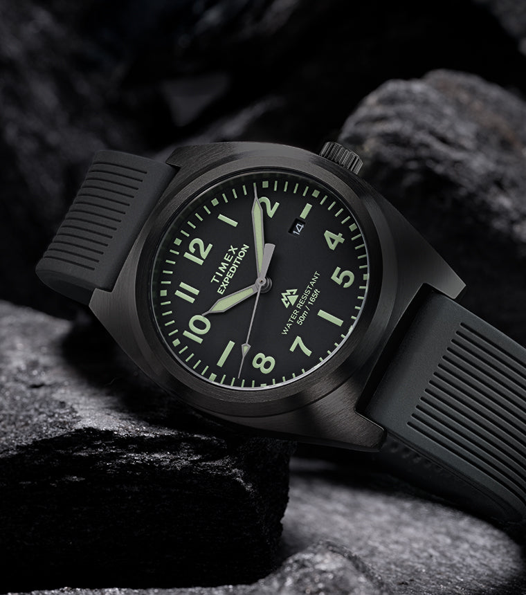 Timex Expedition® Capstone watch with black dial and black silicone strap resting among rugged dark rocks. The watch face glows softly with luminous markers, highlighting its durability, 50-meter water resistance, and adventure-ready design.