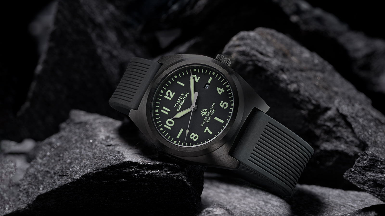 Timex Expedition® Capstone watch with black dial and black silicone strap resting among rugged dark rocks. The watch face glows softly with luminous markers, highlighting its durability, 50-meter water resistance, and adventure-ready design.