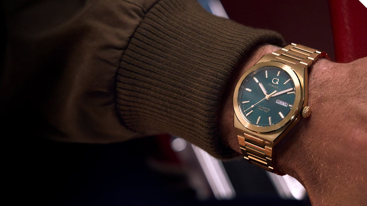 Wrist shot of the gold-tone Q Timex® Continental Day and Date watch with green dial, seen from an angle. The brushed bracelet and bold dial stand out in a relaxed setting, reflecting a retro-inspired luxury look.