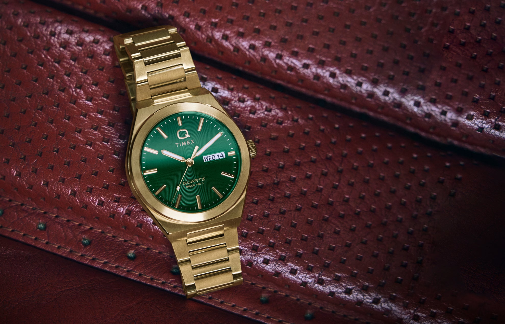 A gold-tone Q Timex® Continental Day and Date watch with a vibrant green dial rests on a red perforated leather car seat. The watch features a brushed stainless-steel bracelet, polished bezel, and a day-date display at 3 o’clock, combining bold color with vintage-inspired design.