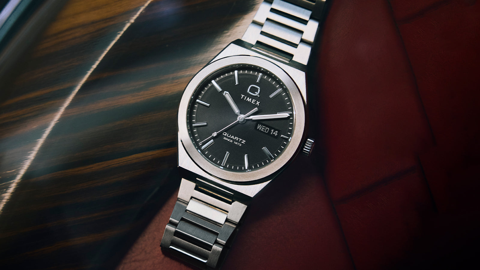 The Q Timex® Continental Day and Date watch lies on a red leather and wood dashboard. Its circular black dial, polished bezel, and linear indices highlight Timex’s modern take on a retro quartz classic.