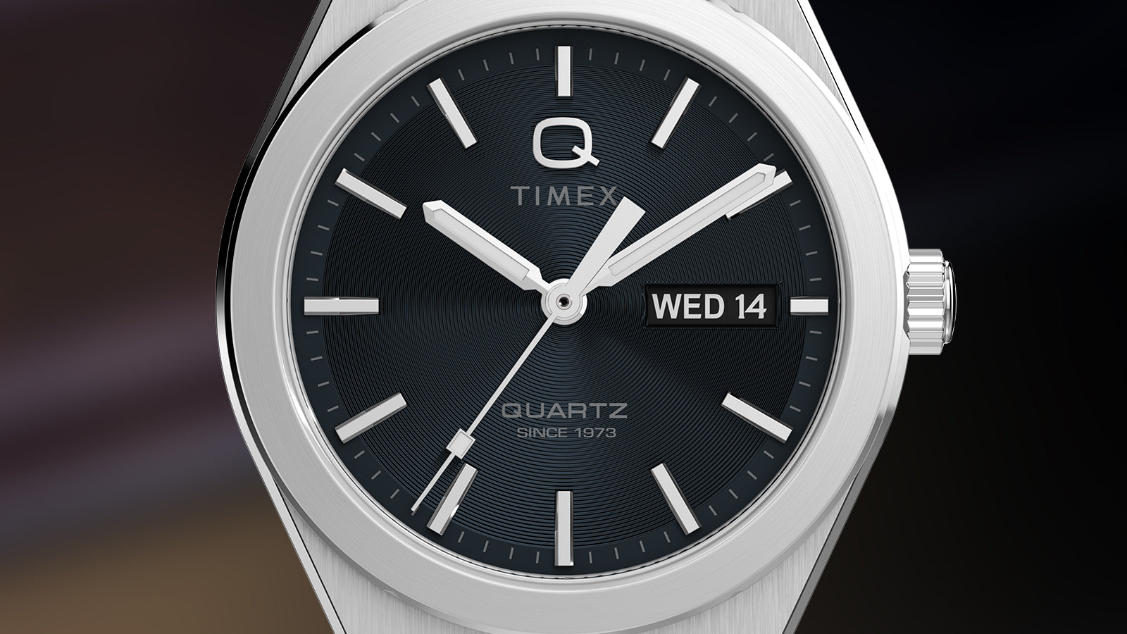 Macro close-up of the Q Timex® Continental Day and Date dial, revealing concentric texturing, luminous hands, and the “Q Timex” logo. The date window at 3 o’clock and brushed bezel emphasize craftsmanship and clarity