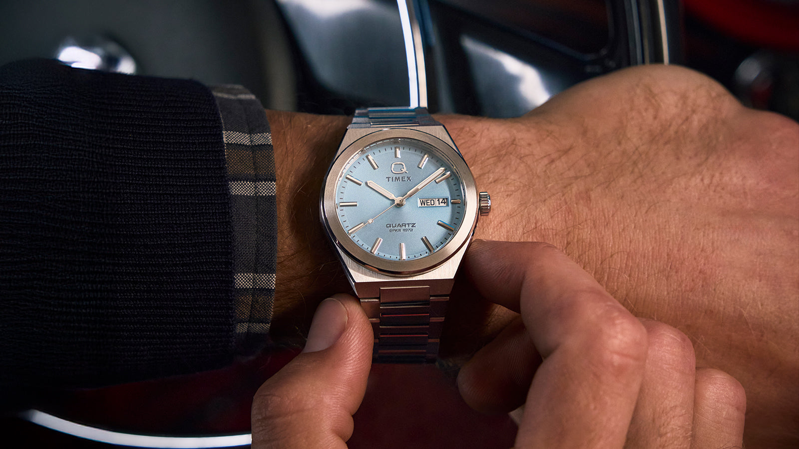 Wrist shot of the Q Timex® Continental Day and Date with an ice-blue dial, worn under a dark cuff with plaid lining. The stainless-steel bracelet and pale blue face offer a cool, sophisticated look that blends classic and contemporary design.