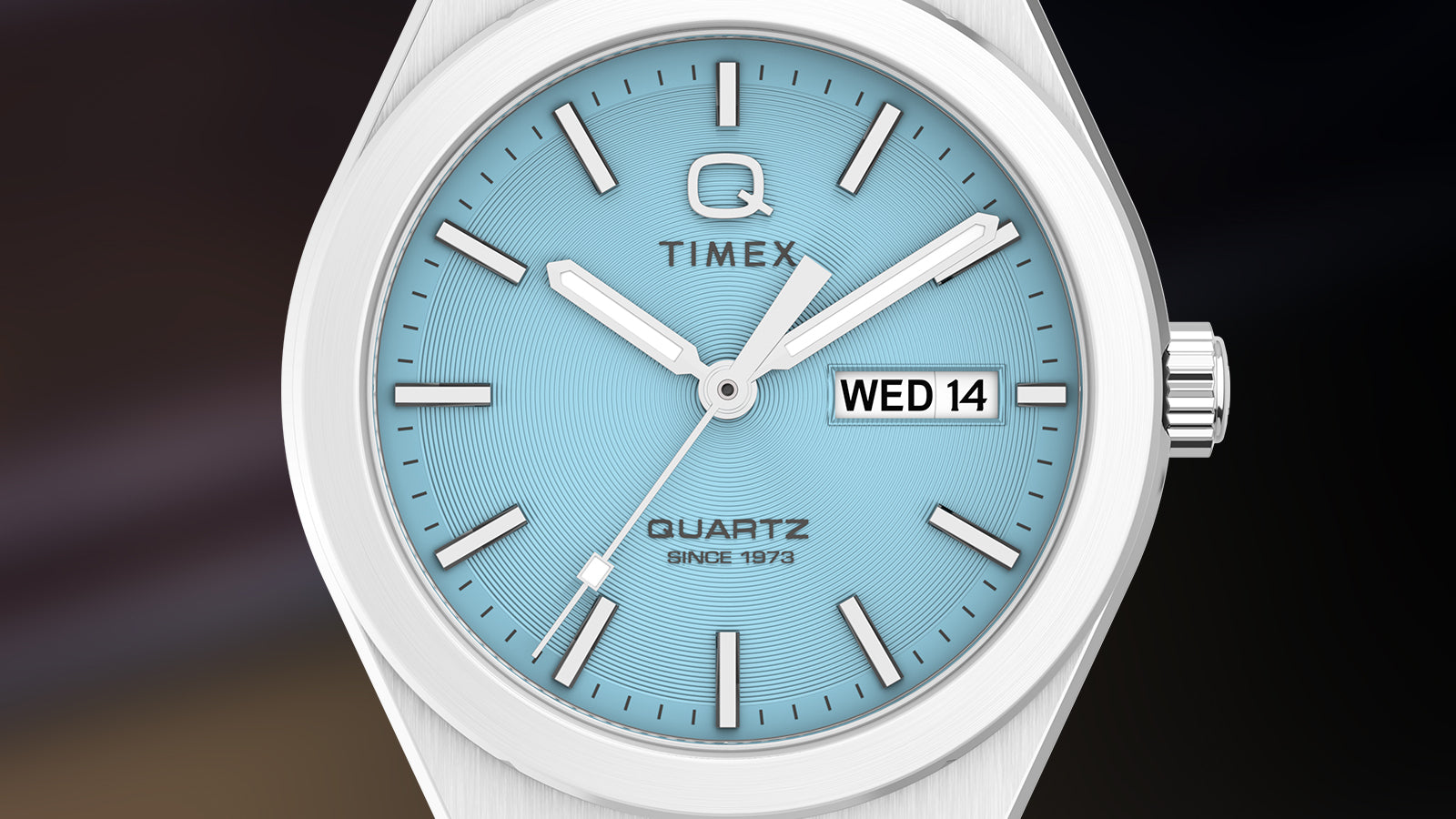 Macro close-up of the Q Timex® Continental Day and Date ice-blue dial. The image highlights concentric texture detailing, silver-tone hour markers, and the day-date display at 3 o’clock, showcasing precision and clarity.