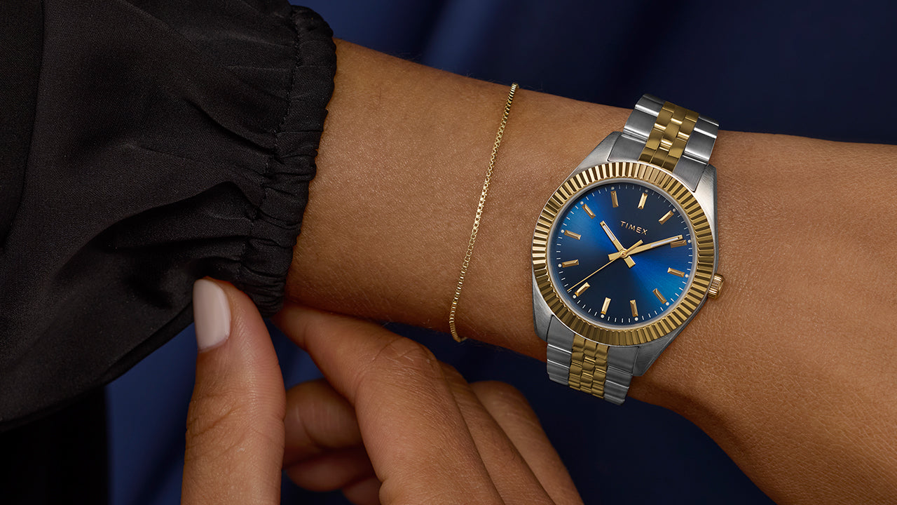 Timex Legacy watch worn on wrist with two-tone bracelet and blue dial, styled with a delicate gold bracelet and dark sleeve for a refined finish.