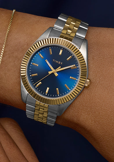 Timex Legacy watch worn on wrist with two-tone bracelet and blue dial, styled with a delicate gold bracelet and dark sleeve for a refined finish.