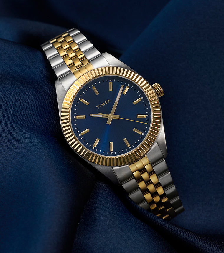 Timex Legacy watch with a blue dial, gold-tone markers, and a two-tone stainless-steel bracelet, displayed on deep blue satin fabric for an elegant look.