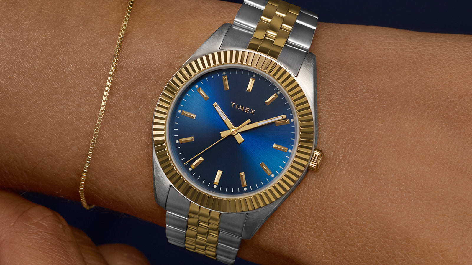 Timex Legacy watch worn on wrist with two-tone bracelet and blue dial, styled with a delicate gold bracelet and dark sleeve for a refined finish.