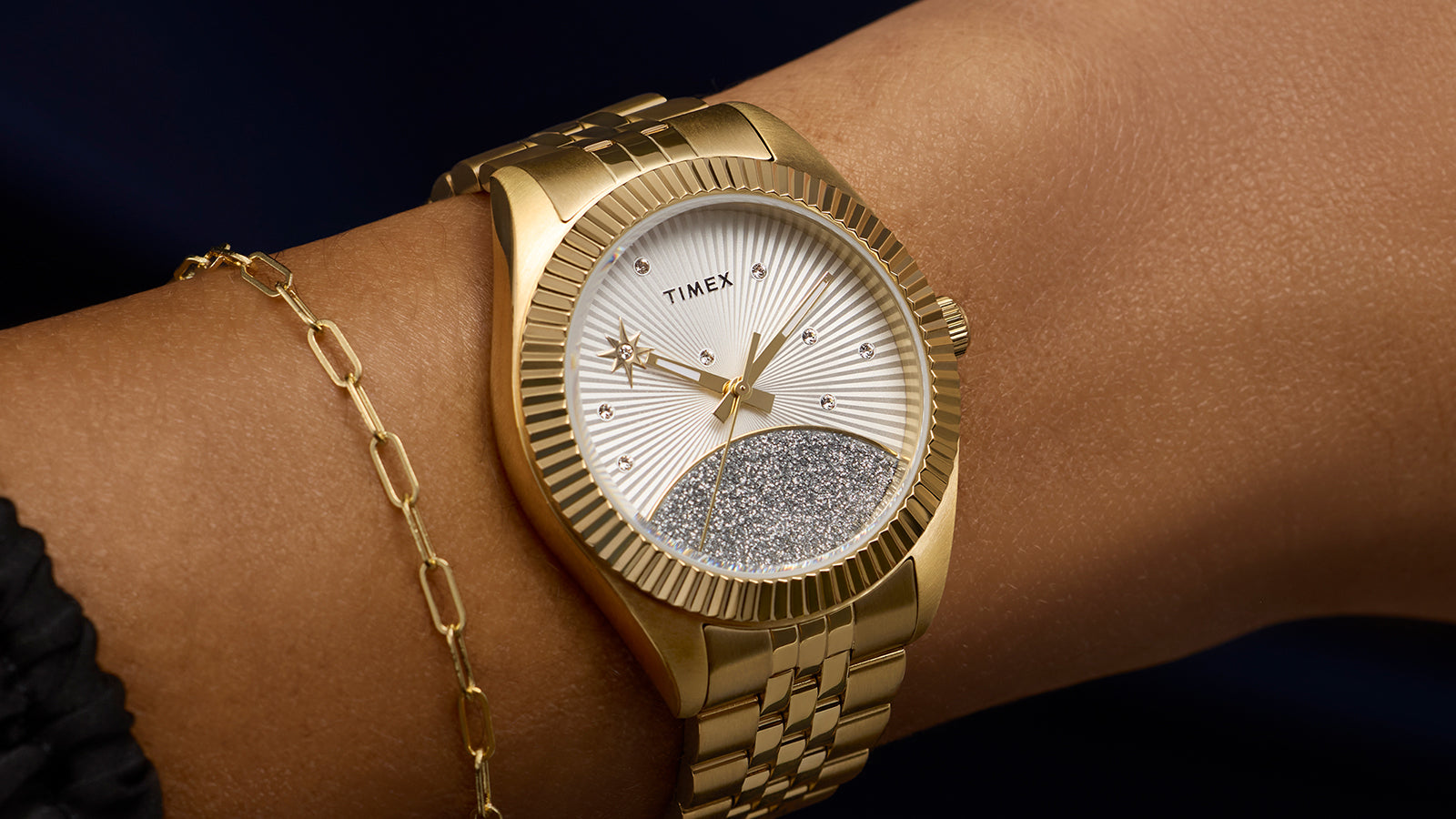 Timex Legacy watch in gold-tone stainless steel with a silver sunray dial featuring crystal accents and a glitter-textured lower half, worn on wrist with a delicate gold bracelet.