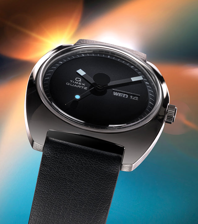 Angled view of the Q Timex® 1975 Enigma watch with a black dial, polished stainless-steel case, and black leather strap set against a glowing space-inspired background.