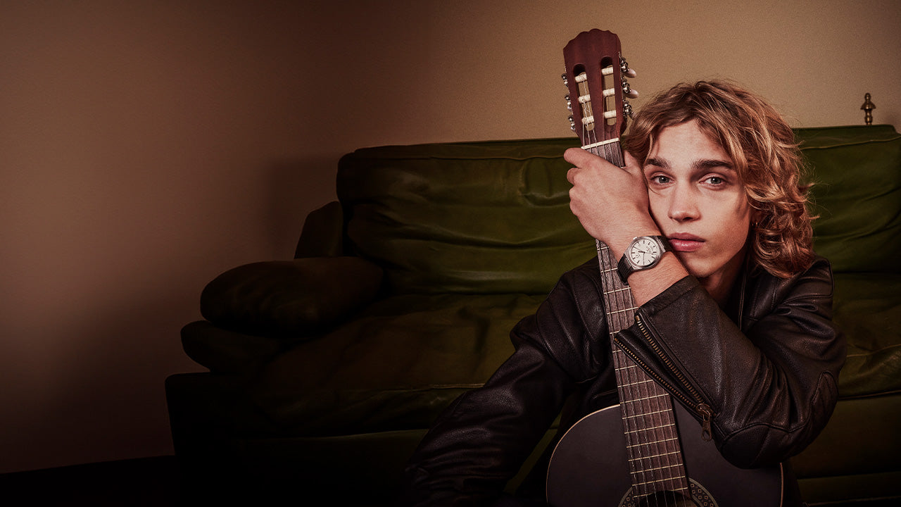 Model seated on a green sofa, embracing an acoustic guitar while wearing the Q Timex® Continental GMT watch, styled in a black leather jacket for a relaxed, vintage-inspired look.