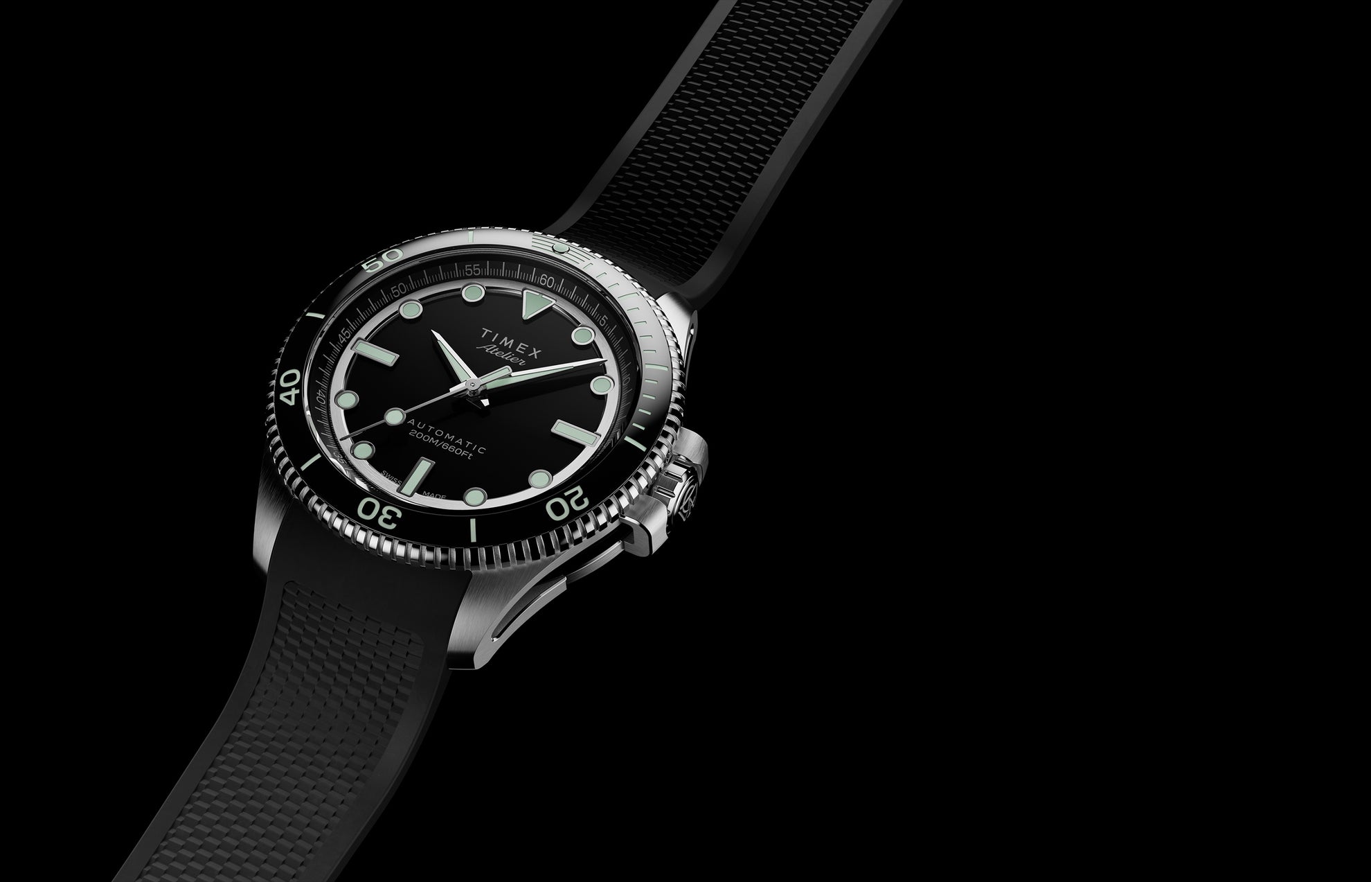 Timex Atelier automatic dive watch with stainless steel case, black dial, luminous markers, and black textured rubber strap on a dark background.