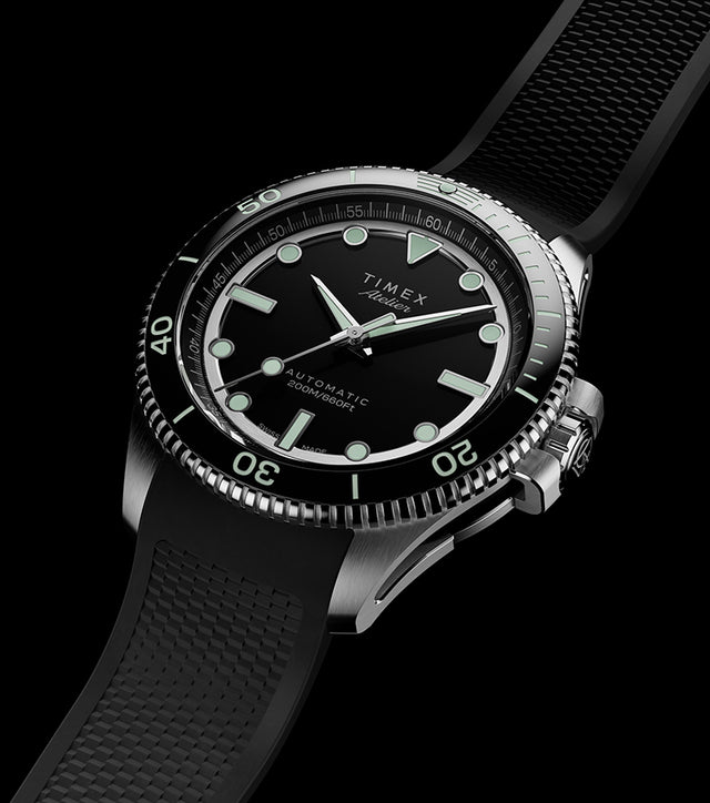 Timex Atelier automatic dive watch with stainless steel case, black dial, luminous markers, and black textured rubber strap on a dark background.
