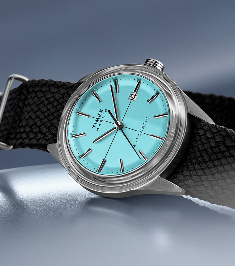 Timex Marlin Jet Automatic with aqua blue dial, stainless steel case, and black woven strap displayed on a metallic blue surface.