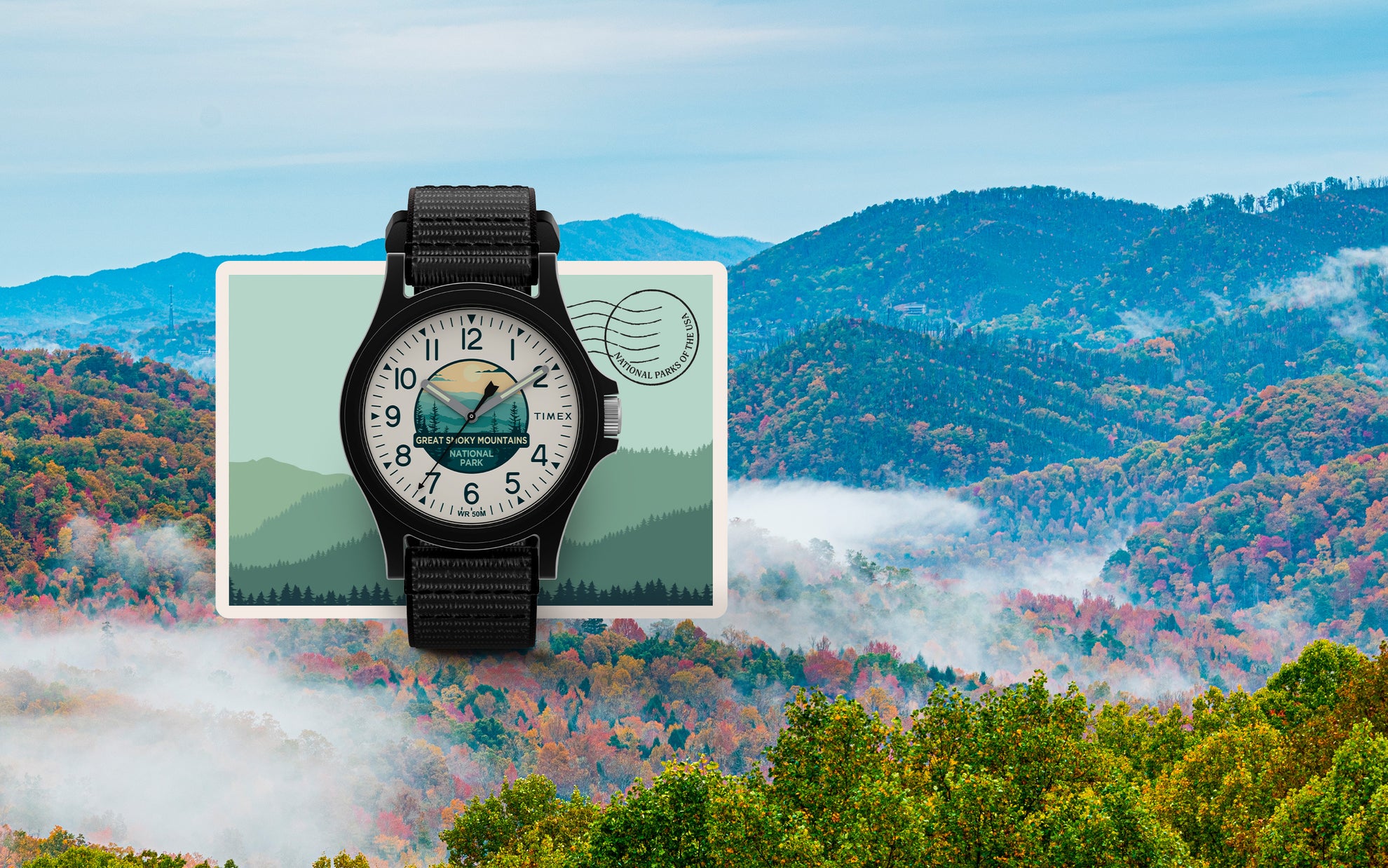 Timex x Great Smoky Mountains National Park watch displayed over a scenic landscape of the Smoky Mountains in fall, with rolling mist and colorful foliage. The watch has a black case, black fabric strap, and a dial illustrated with a mountain sunrise, trees, and a bear silhouette. 'Great Smoky Mountains National Park' is printed on the dial.