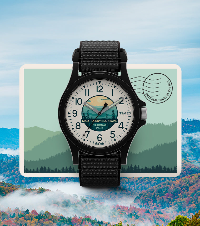 Timex x Great Smoky Mountains National Park watch centered over a stylized national park postcard background, featuring layered tree silhouettes and a circular postmark labeled 'National Parks of the USA'. The watch design includes a black case, black fabric strap, and an illustrated nature scene on the dial.