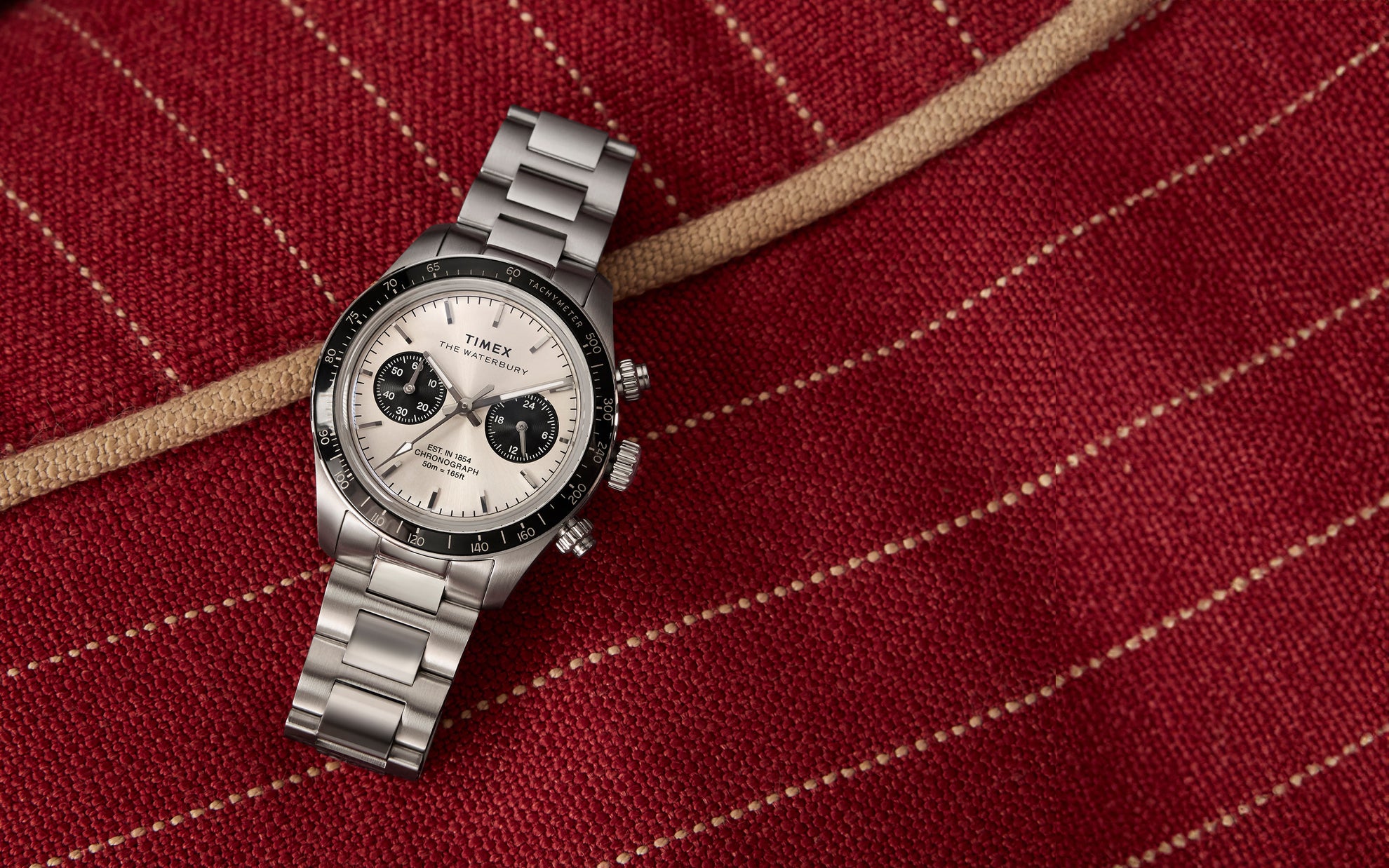 A detailed shot of the Timex Waterbury Heritage Chronograph watch resting on a rich, textured red fabric with beige stitching. The stainless steel bracelet, black tachymeter bezel, and silver dial with contrasting black subdials are prominently displayed, highlighting the watch’s refined yet sporty design.