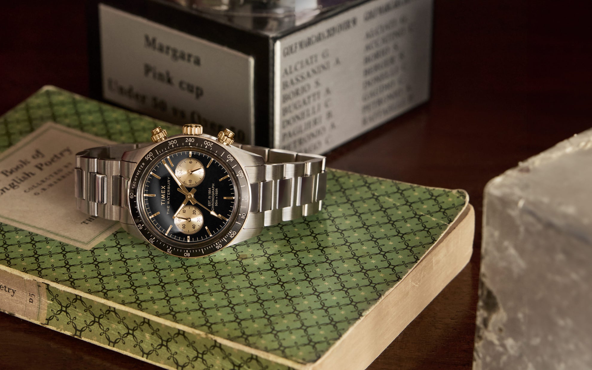 A close-up shot of the Timex Waterbury Heritage Chronograph resting on a vintage green-patterned book. The stainless steel bracelet, black dial with gold subdials, and gold-tone pushers and crown stand out, evoking a classic and refined aesthetic.
