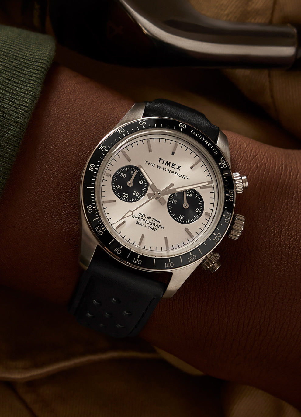 Close-up of a Timex Waterbury Heritage Chronograph watch on a wrist, with a silver dial, black subdials, and a black leather strap, emphasizing its sporty elegance.