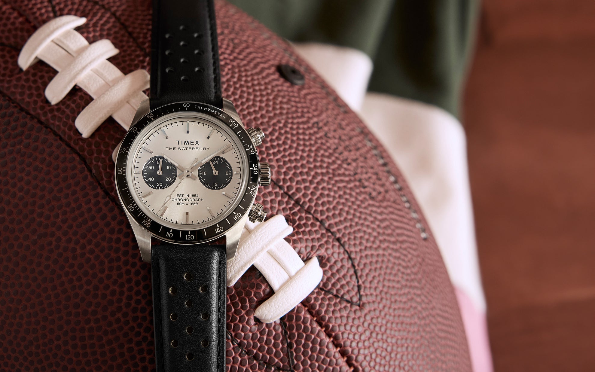 A Timex Waterbury Heritage Chronograph watch with a black perforated leather strap, resting on a football, featuring a silver dial with black subdials and a tachymeter bezel.