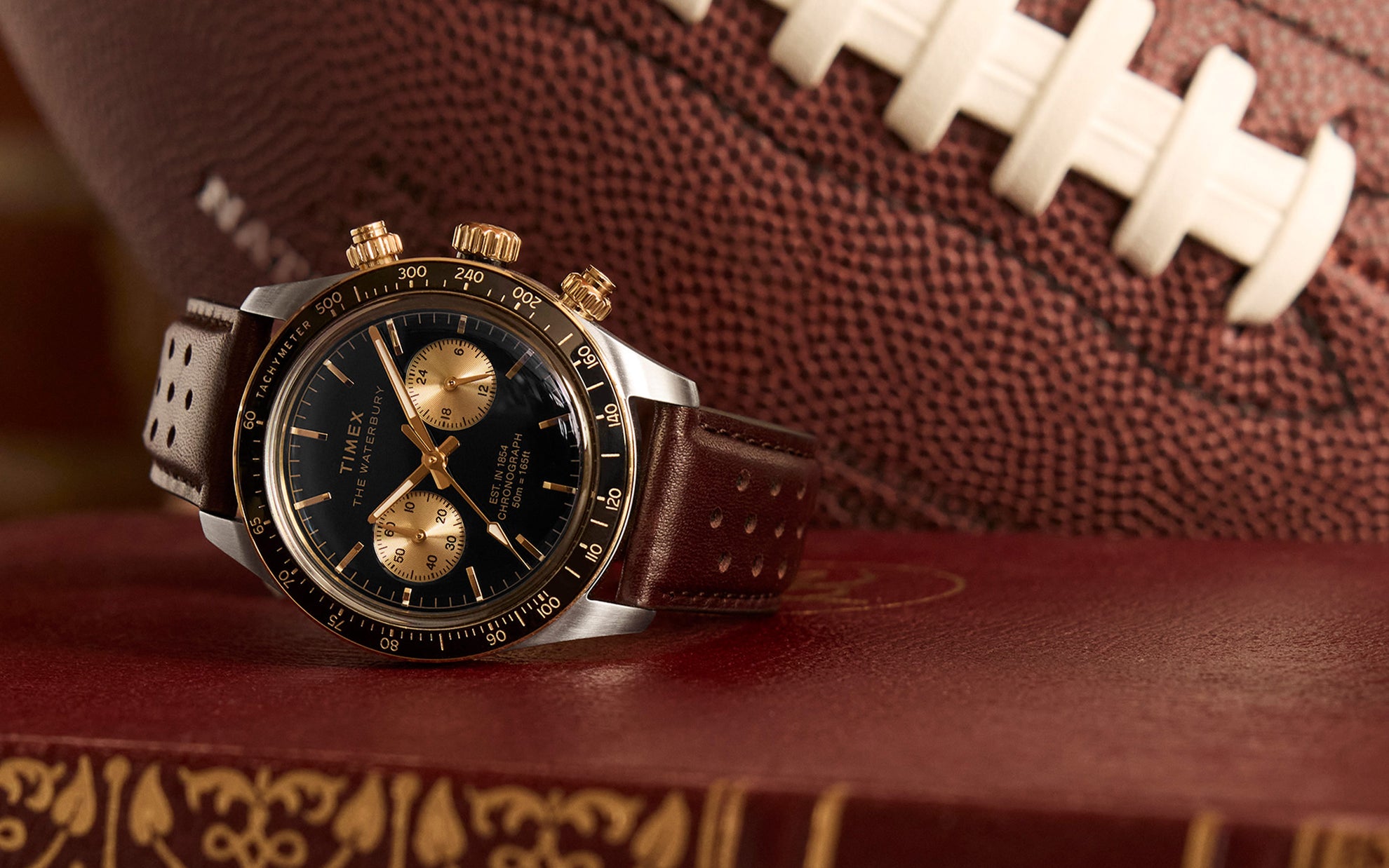A close-up shot of the Timex Waterbury Heritage Chronograph resting on a vintage leather-bound book, with an American football in the background. The black dial with gold-tone accents, perforated brown leather strap, and polished case create a classic sports-inspired aesthetic.