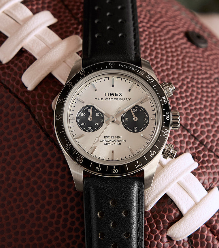 A Timex Waterbury Heritage Chronograph watch with a black perforated leather strap, resting on a football, featuring a silver dial with black subdials and a tachymeter bezel.