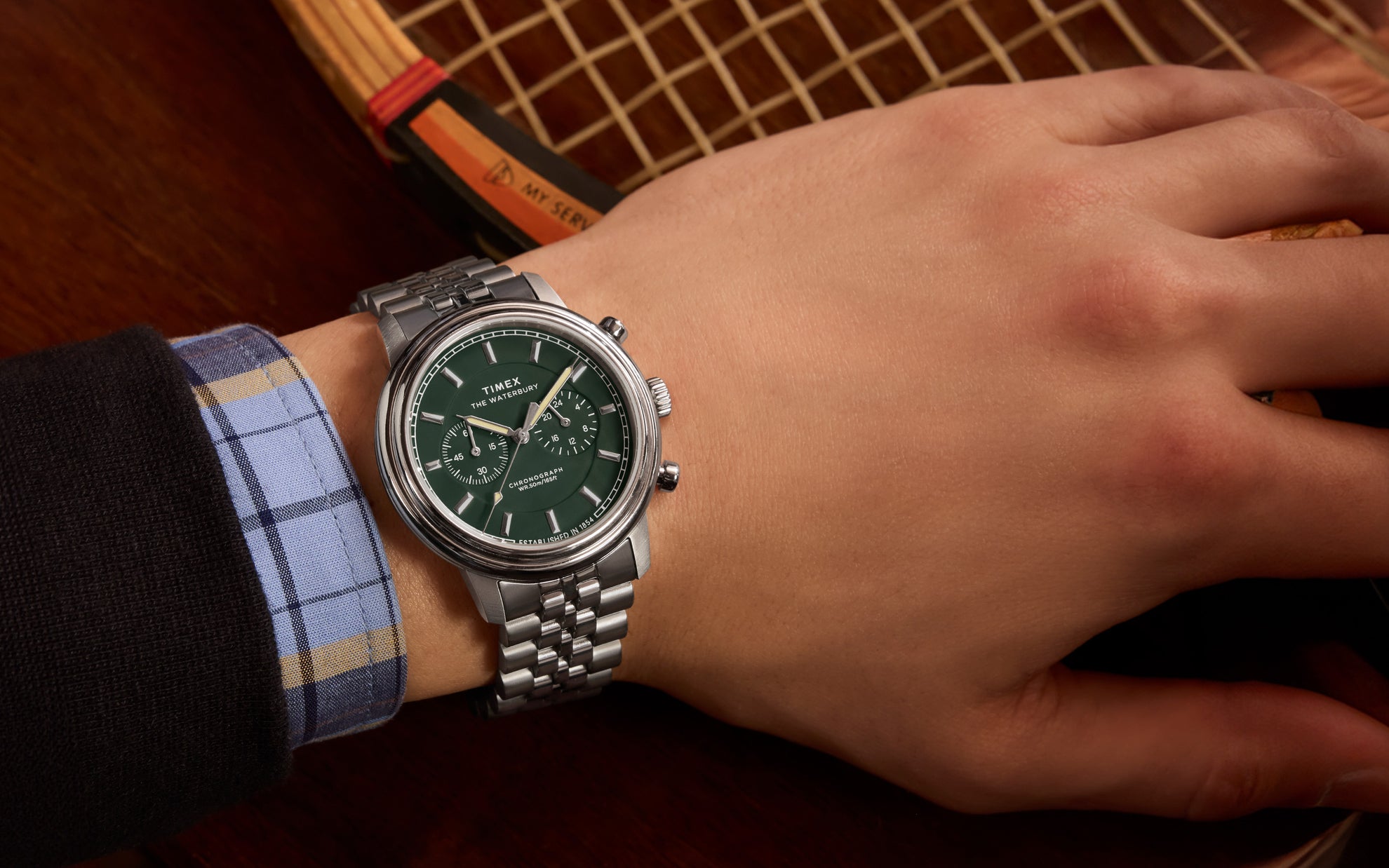 A Timex Waterbury Metropolitan Chronograph watch with a green dial, silver-tone markers, and a stainless steel bracelet, worn on a person’s wrist with a vintage wooden tennis racket in the background.