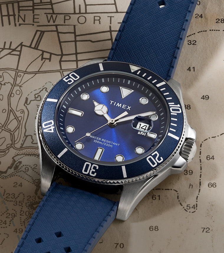 A Timex Harborside Coast watch with a navy blue silicone strap rests on a nautical chart map, showing the coast near Newport. The stainless steel case and blue dial reflect soft light, and the watch face reads “Water Resistant 100m / 330ft” with a magnified date window at 3 o'clock.