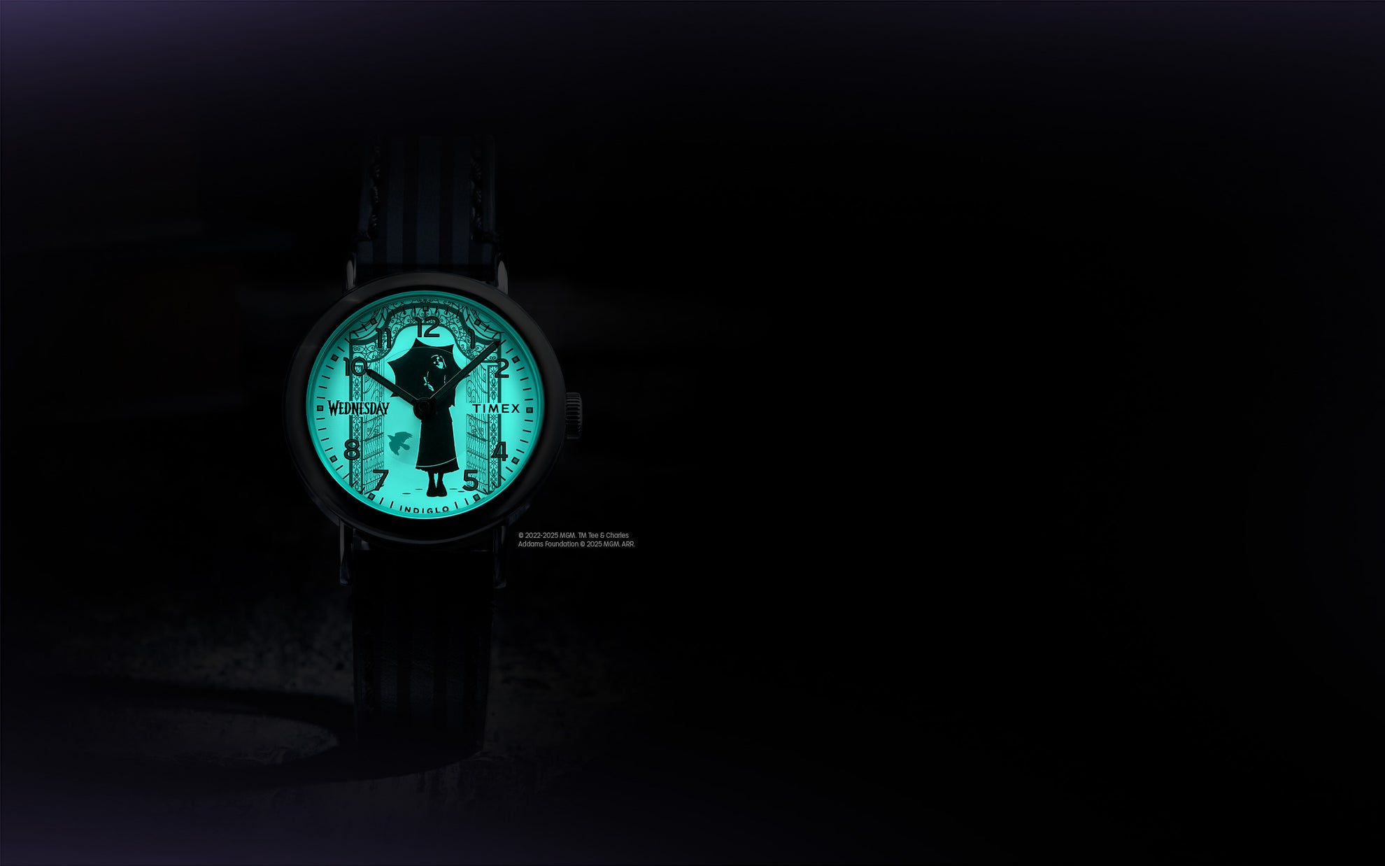 Flatlay of the Timex x Wednesday watch with its Indiglo backlight glowing green in the dark, revealing gothic gate artwork around the dial.