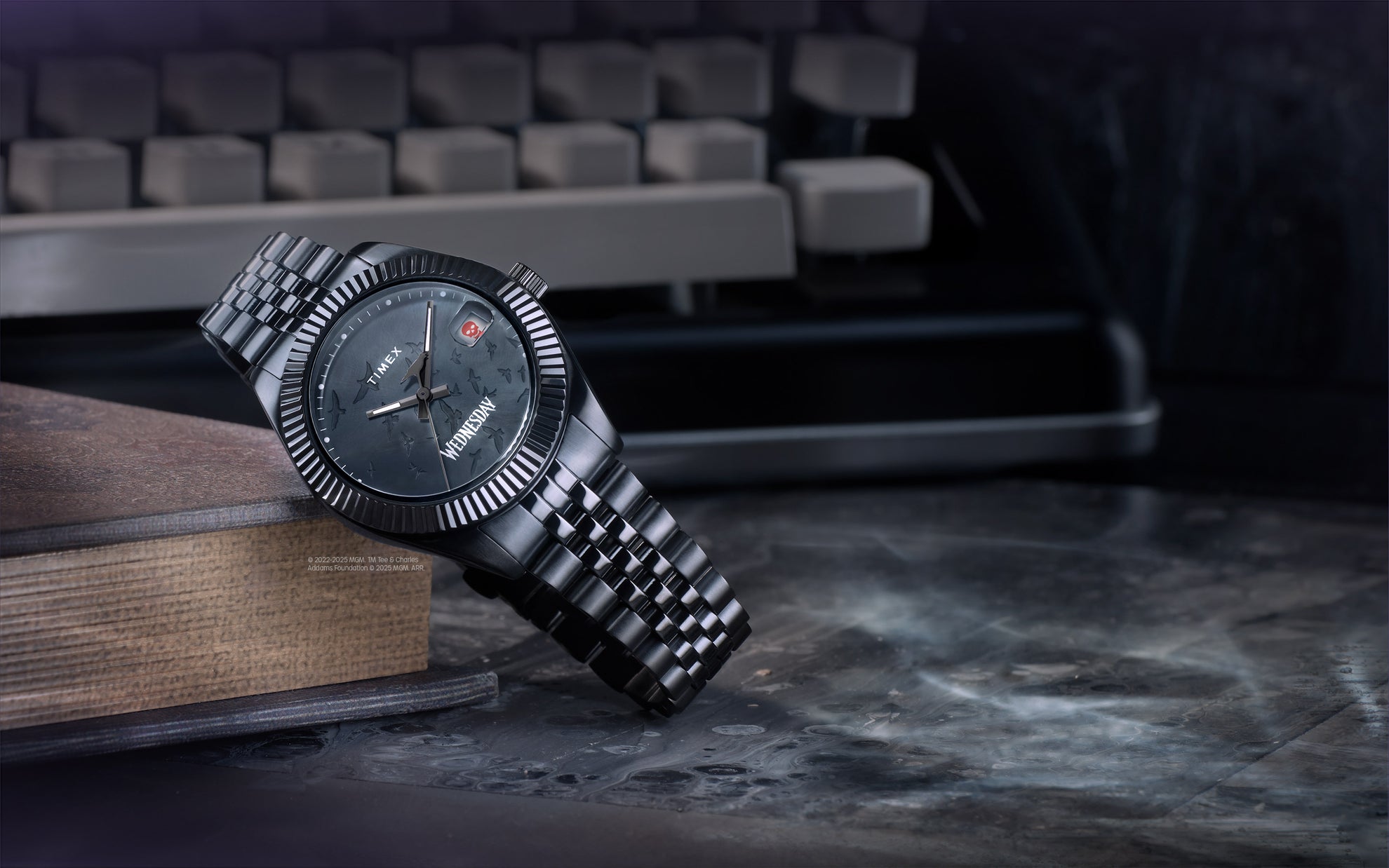 The Timex x Wednesday Legacy watch resting atop a stack of old books, with a vintage typewriter in the background. The watch features a black stainless-steel case and bracelet, a dark textured dial with scattered raven silhouettes, a skull icon at the date window, and “Wednesday” printed at the 6 o’clock position.